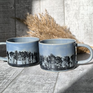 Op de afbeelding: Twee blauwe keramische mokken met een zwart silhouet van een bos scène. De mokken staan op een wit houten oppervlak.
