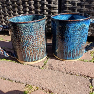 May include: Two blue ceramic mugs with a rope-like handle. The mugs have a textured, mottled glaze with brown and blue tones.