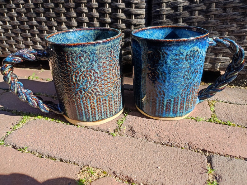 May include: Two blue ceramic mugs with a rope-like handle. The mugs have a textured, mottled glaze with brown and blue tones.