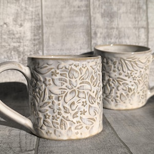 May include: Two off-white ceramic mugs with embossed floral designs. The mugs have a cream-colored glaze and curved handles. The floral pattern includes leaves, flowers, and other botanical elements. The mugs are set on a gray wooden surface.
