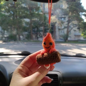 May include: A crocheted orange and brown fire character with a smiling face, holding a brown log, hanging from a rearview mirror in a car.