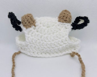 May include: White crocheted cow hat with brown and black ears. The hat has a brown string tie.
