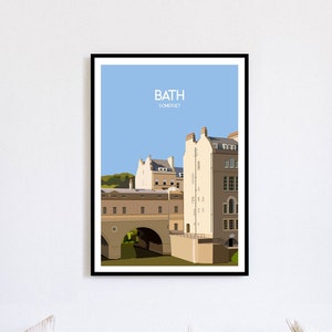 Puede incluir: Una impresión artística enmarcada que presenta una ilustración estilizada de Bath, Somerset. La obra de arte representa edificios y un puente bajo un cielo azul, con el texto "BATH SOMERSET" en la parte superior. La impresión se muestra en una pared blanca sobre un sofá blanco.