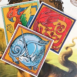 Può includere: Tre adesivi quadrati con illustrazioni di creature mitiche. Uno mostra un grifone rosso con ali gialle e verdi, un altro un drago rosso e il terzo un narvalo blu. Ognuno ha uno sfondo giallo e un bordo decorativo.