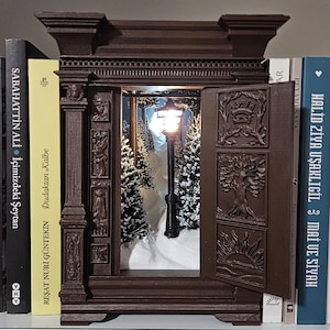 Puede incluir: Un mueble en miniatura marrón oscuro, ornamentado, con puertas abiertas que revelan una escena invernal con una farola y árboles cubiertos de nieve. El mueble está flanqueado por libros en una estantería, creando una exhibición decorativa.