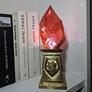 Peut inclure: Une lampe en forme de cristal rouge à surface texturée est posée sur un piédestal doré. Le piédestal présente un motif de bouclier avec un emblème de lion. La lampe est placée sur une étagère blanche à côté d'une rangée de livres.