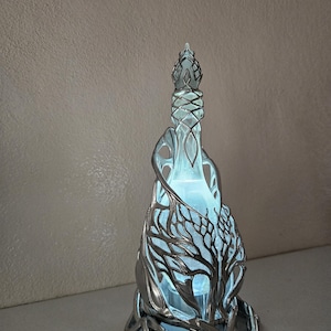May include: A decorative glass bottle with a silver metal tree design. The bottle has a unique shape and is illuminated with a soft blue light, creating a mystical ambiance. The tree design wraps around the bottle, adding an artistic touch.