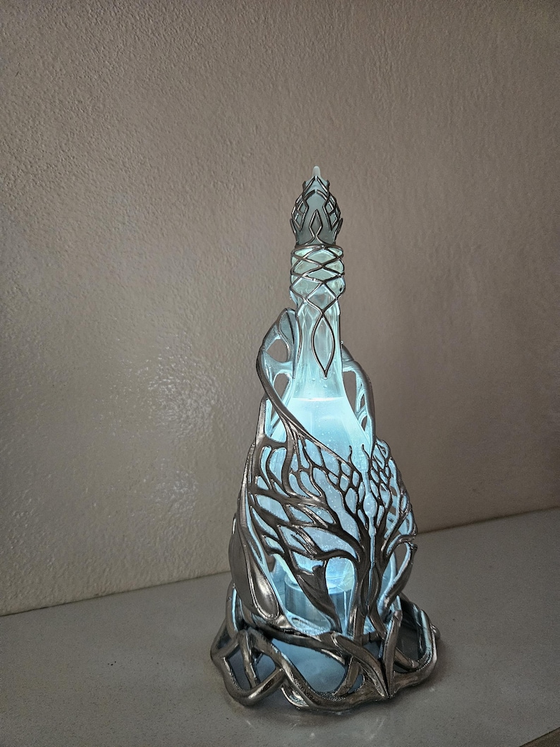 May include: A decorative glass bottle with a silver metal tree design. The bottle has a unique shape and is illuminated with a soft blue light, creating a mystical ambiance. The tree design wraps around the bottle, adding an artistic touch.