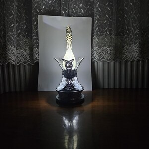 May include: A decorative bottle with an ornate black base and a detailed, openwork design. The bottle has a slender, elongated shape with a light-colored interior, and is topped with a decorative stopper. The bottle is displayed on a dark wooden surface.