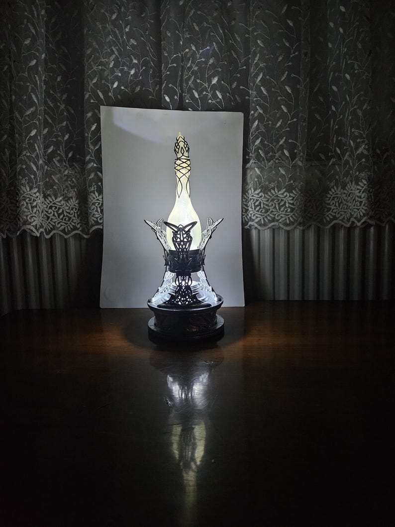 May include: A decorative bottle with an ornate black base and a detailed, openwork design. The bottle has a slender, elongated shape with a light-colored interior, and is topped with a decorative stopper. The bottle is displayed on a dark wooden surface.