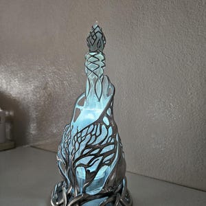 May include: A decorative glass bottle with a silver-coloured, tree-like design. The bottle has a blue interior that glows, and the silver design wraps around the glass. The bottle has a decorative stopper.