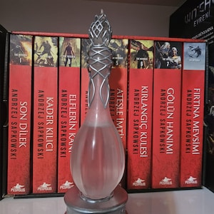May include: A decorative glass bottle with a silver-coloured metal base and accents, standing in front of a row of red books. The bottle has a teardrop shape and a detailed top. The books are part of a series.