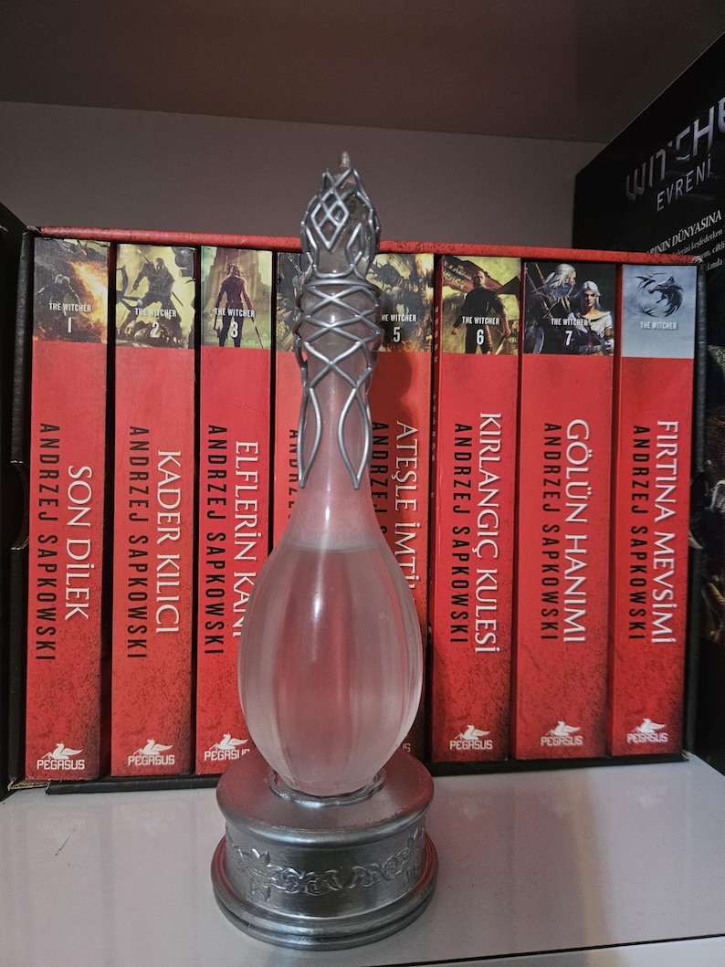 May include: A decorative glass bottle with a silver-coloured metal base and accents, standing in front of a row of red books. The bottle has a teardrop shape and a detailed top. The books are part of a series.