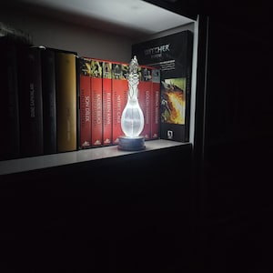 May include: A decorative glass bottle with an intricate design, lit up by a soft light. The bottle is on a dark base, displayed on a shelf with a collection of books. The books have red and yellow spines.