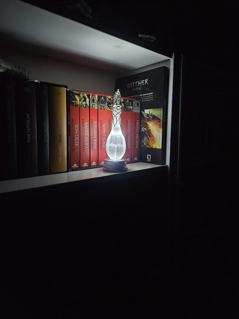 May include: A decorative glass bottle with an intricate design, lit up by a soft light. The bottle is on a dark base, displayed on a shelf with a collection of books. The books have red and yellow spines.