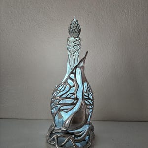 May include: An ornate glass bottle with a silver metal filigree design. The bottle features a long neck and a decorative stopper. The interior is illuminated with a soft blue light. The bottle's design is artistic and elegant, suitable for display.