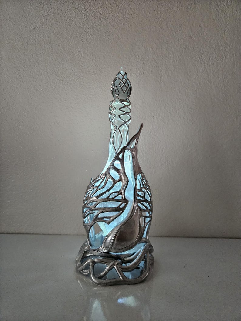 May include: An ornate glass bottle with a silver metal filigree design. The bottle features a long neck and a decorative stopper. The interior is illuminated with a soft blue light. The bottle's design is artistic and elegant, suitable for display.
