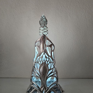 May include: An ornate, silver-toned glass bottle with a detailed, openwork design. The bottle has a light blue interior that illuminates the intricate patterns. The stopper is also silver and features a geometric design. The bottle is approximately 25 cm tall.