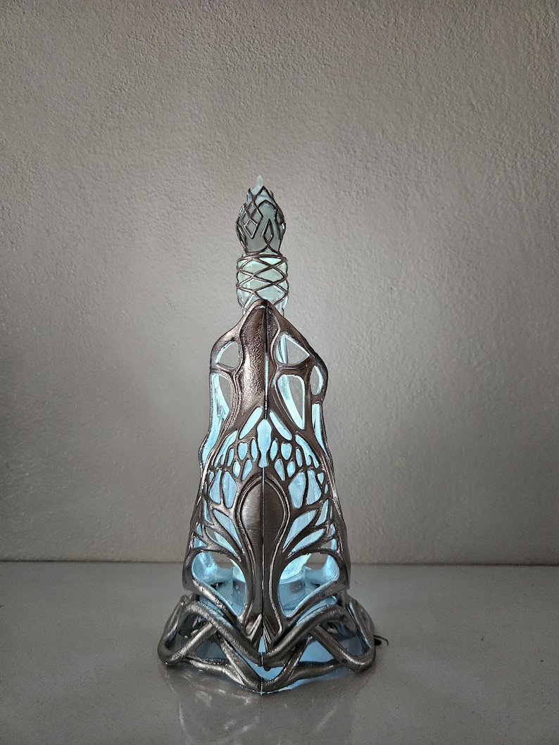 May include: An ornate, silver-toned glass bottle with a detailed, openwork design. The bottle has a light blue interior that illuminates the intricate patterns. The stopper is also silver and features a geometric design. The bottle is approximately 25 cm tall.