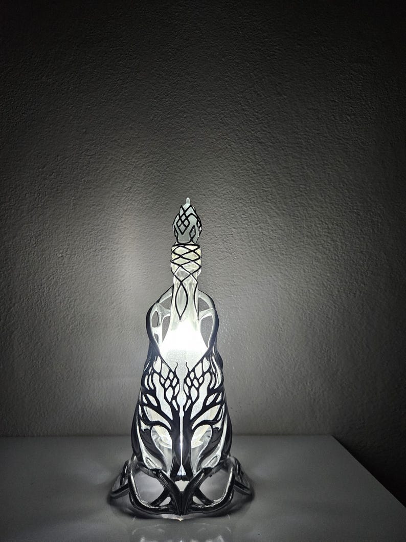 May include: A decorative lamp with a black and white tree design. The lamp has an openwork pattern and a glowing interior. The base and top are also intricately designed. The lamp is lit, creating a soft, ambient glow.