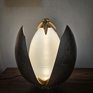 May include: A unique, illuminated table lamp with a floral design. The lamp features a central, egg-shaped light source encased in stylized, petal-like structures. The petals are a dark gray with etched details, and the light source glows with a soft, warm light.