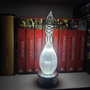 May include: An illuminated glass sculpture with a bulbous body and a detailed, patterned top. The sculpture is set on a dark, ornate base. The background features a row of books with red spines.