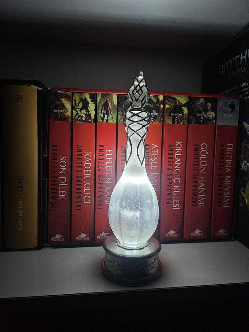 May include: An illuminated glass sculpture with a bulbous body and a detailed, patterned top. The sculpture is set on a dark, ornate base. The background features a row of books with red spines.