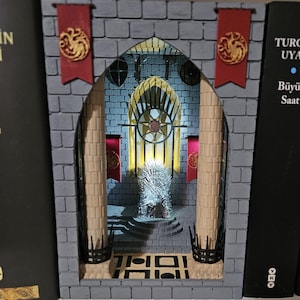 Puede incluir: Un separador de libros de ladrillo gris con una escena en miniatura del Trono de Hierro de Juego de Tronos. El trono está iluminado con una luz blanca cálida. El separador de libros tiene una pancarta roja y dorada con un emblema de dragón a cada lado.