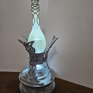 May include: A decorative lamp with a glowing, light green, teardrop-shaped glass bottle. The bottle is held in place by an ornate silver-colored metal stand with intricate designs. The lamp has a decorative top and base, and the light emanates from within the bottle.