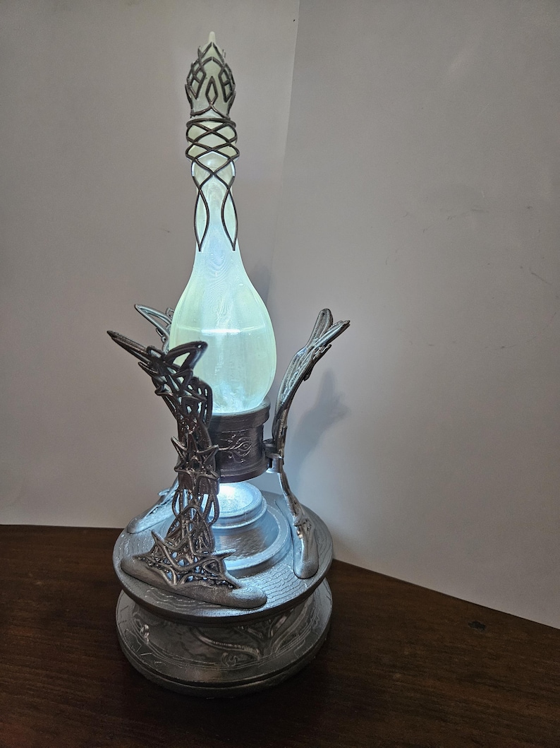 May include: A decorative lamp with a glowing, light green, teardrop-shaped glass bottle. The bottle is held in place by an ornate silver-colored metal stand with intricate designs. The lamp has a decorative top and base, and the light emanates from within the bottle.