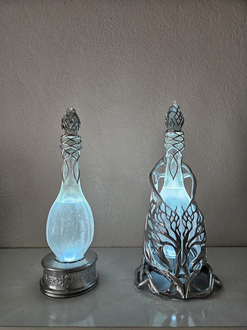 May include: Two decorative glass bottles with silver detailing and a soft blue glow. One has a simple design, the other features intricate tree-like metalwork. Both are illuminated from within, creating a magical ambiance.
