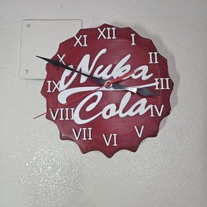 May include: A red and white wall clock shaped like a bottle cap, featuring the text "Nuka Cola" in white script. The clock has black hands and Roman numeral hour markers. The clock is mounted on a white wall.