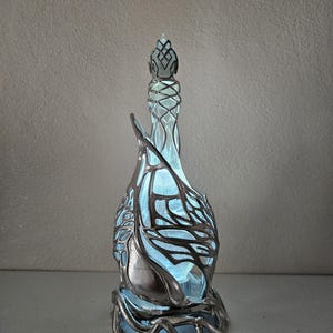 May include: A decorative glass bottle with a distinctive design. The bottle has a light blue interior and is encased in a silver-coloured metal filigree. The filigree has an intricate, organic pattern. The bottle has a tall, slender neck and a decorative stopper.
