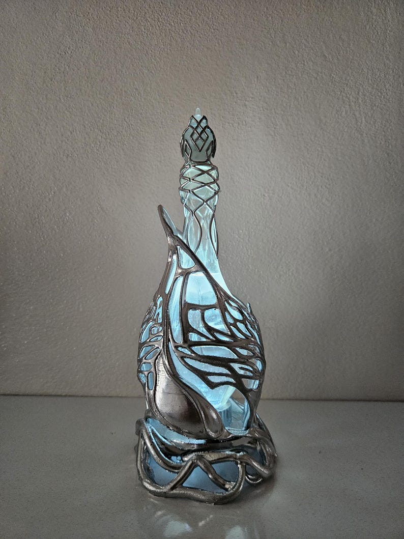 May include: A decorative glass bottle with a distinctive design. The bottle has a light blue interior and is encased in a silver-coloured metal filigree. The filigree has an intricate, organic pattern. The bottle has a tall, slender neck and a decorative stopper.