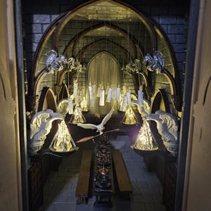 Op de afbeelding: Miniatuur diorama van een gotische eetzaal, met een lange tafel gedekt met miniatuur gerechten en een vliegende uil. De scène wordt verlicht door hangende kaarsen en wandkandelaars, wat een warme sfeer creëert. De muren zijn gemaakt van grijze baksteen.