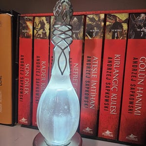 May include: An illuminated glass bottle with a silver metal base and top. The bottle has a distinctive shape and is lit from within, creating a soft glow. The background features a shelf with several books.