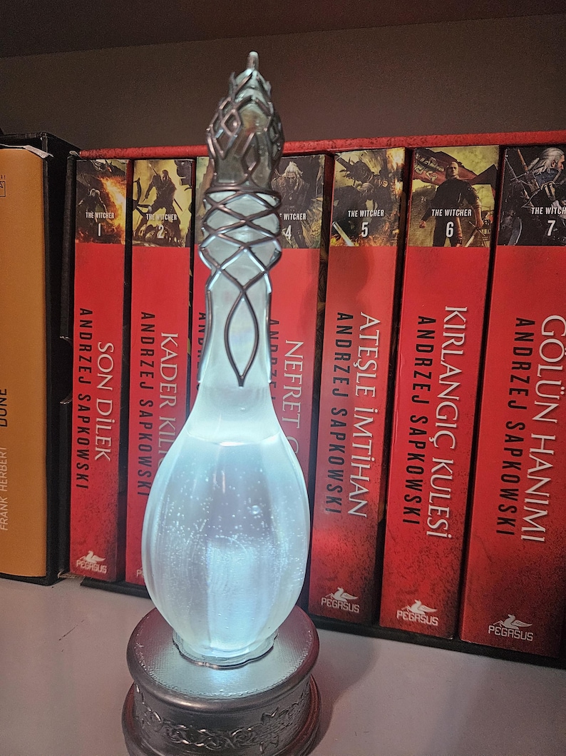May include: An illuminated glass bottle with a silver metal base and top. The bottle has a distinctive shape and is lit from within, creating a soft glow. The background features a shelf with several books.