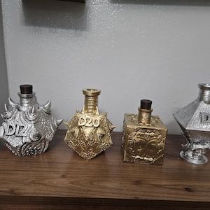 Può includere: Quattro bottiglie decorative nei toni dell'argento e dell'oro, ciascuna con un design geometrico unico. Le bottiglie hanno un marchio D20 e sono esposte su una superficie di legno.