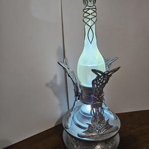 May include: A decorative lamp with a silver base and intricate metalwork. The lamp features a glowing, light green glass bottle-shaped center, and a detailed top. The base has a circular design with ornate patterns, and the lamp is lit from within.