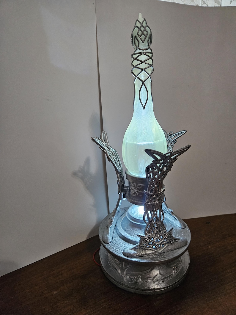 May include: A decorative lamp with a silver base and intricate metalwork. The lamp features a glowing, light green glass bottle-shaped center, and a detailed top. The base has a circular design with ornate patterns, and the lamp is lit from within.