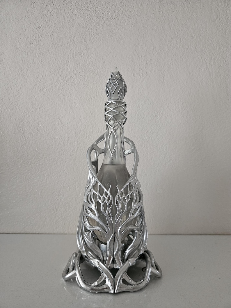 May include: A silver decorative bottle with an elaborate metal filigree design. The bottle has a narrow neck and a detailed stopper. The metalwork features a stylized, organic pattern, creating a unique and artistic appearance.