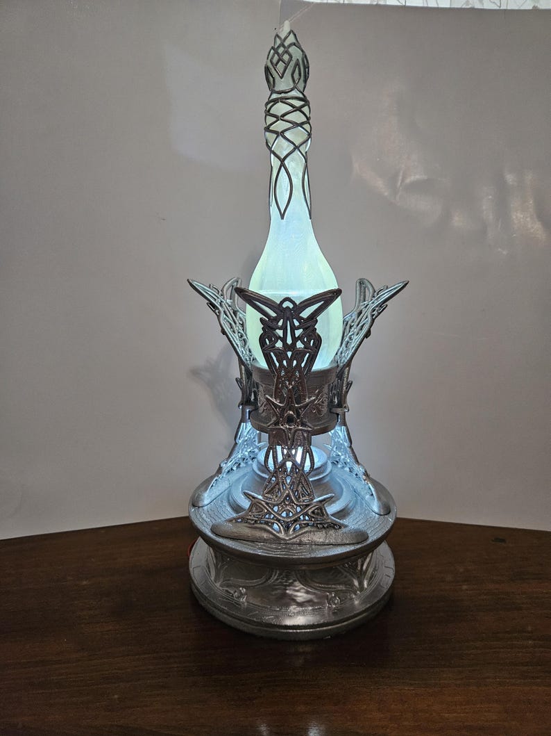 May include: A silver-colored decorative lamp with an intricate design. The lamp features a tall, slender, bottle-shaped center with a glowing blue light. The base and decorative elements are detailed with ornate patterns. The lamp is designed to sit on a flat surface.