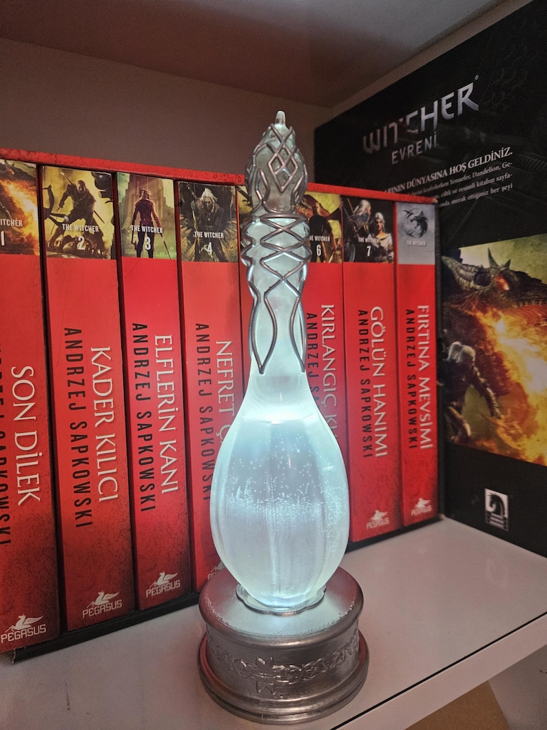 May include: A decorative, glowing glass sculpture with a silver base and intricate detailing. The sculpture is in front of a row of red books with gold lettering. The books are part of a series.