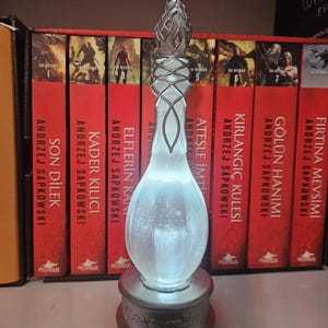 May include: A decorative, illuminated glass bottle with a silver base and intricate metalwork. The bottle has a teardrop shape and a frosted appearance, with a glowing interior. The base is detailed with embossed designs. The background features a row of red books.