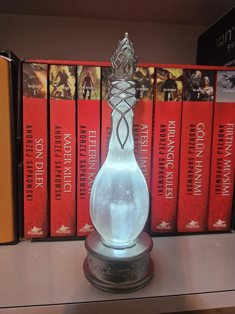 May include: An illuminated glass bottle with a silver base and detailed design. The bottle glows softly from within. Behind it are red books with gold lettering. The books are part of a collection.