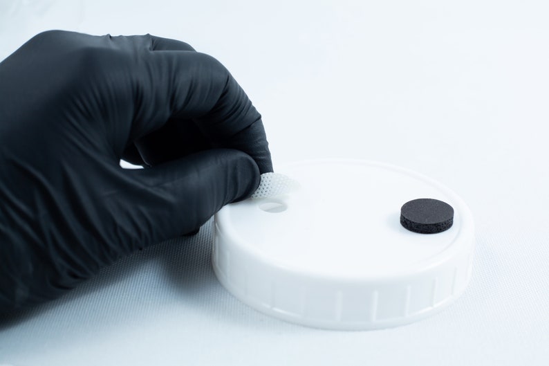 May include: A white plastic lid with a small hole in the center. A gloved hand is holding a white filter over the hole. A black round filter is on the lid.