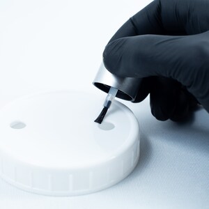 May include: A gloved hand holds a small bottle of nail polish with a black brush over a white plastic lid with two holes.