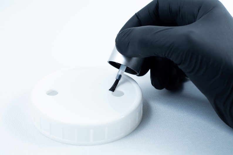May include: A gloved hand holds a small bottle of nail polish with a black brush over a white plastic lid with two holes.