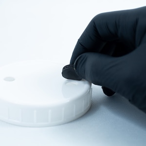 May include: A gloved hand places a black round lid on a white lid with a small hole in the center.