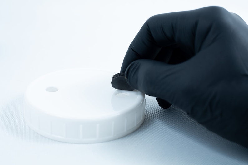 May include: A gloved hand places a black round lid on a white lid with a small hole in the center.